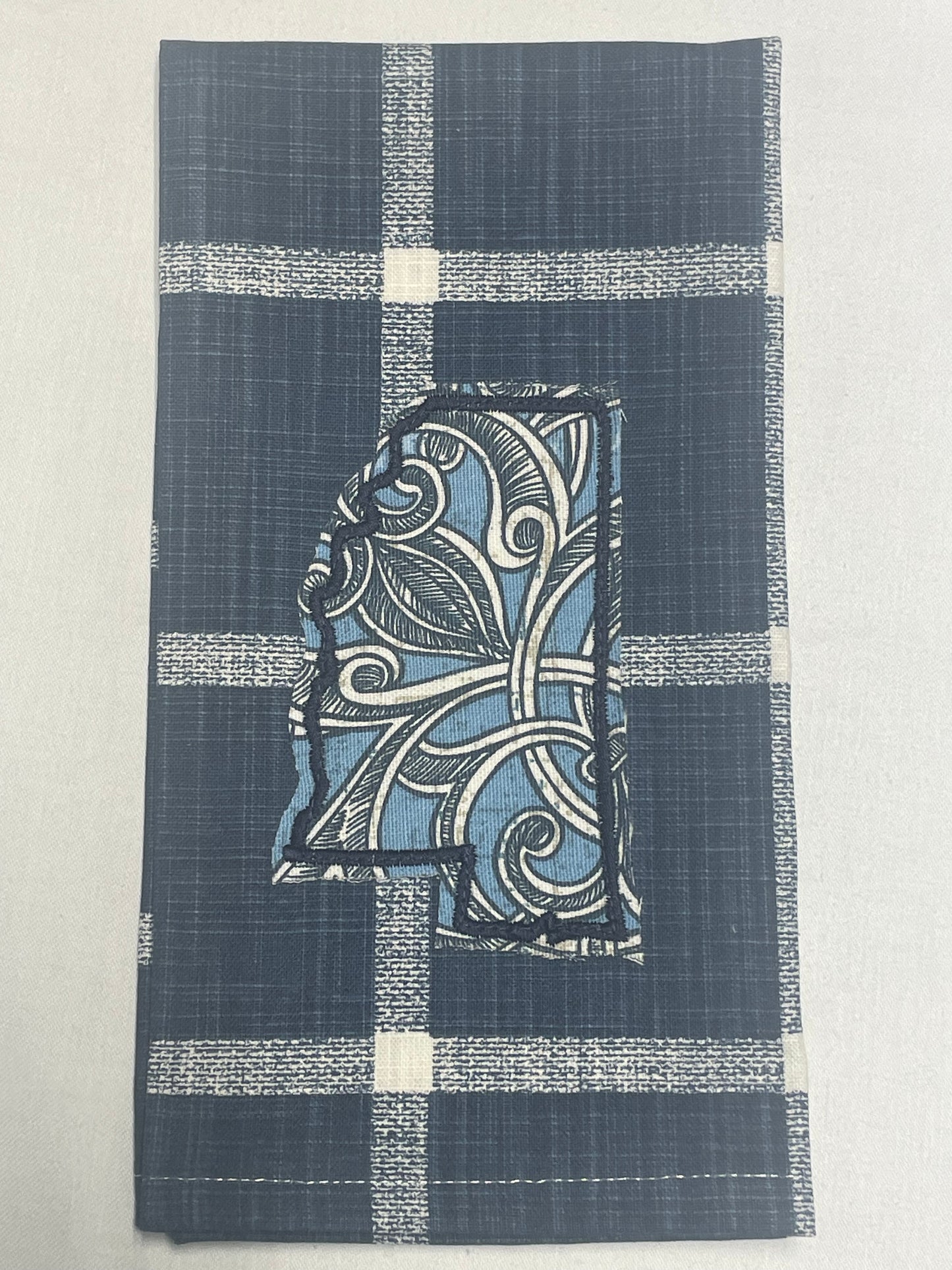 Decorative hand towel featuring embroidered Mississippi state outline with blue and white floral scroll pattern on dark blue plaid background, perfect for southern home decor and state pride gifts.