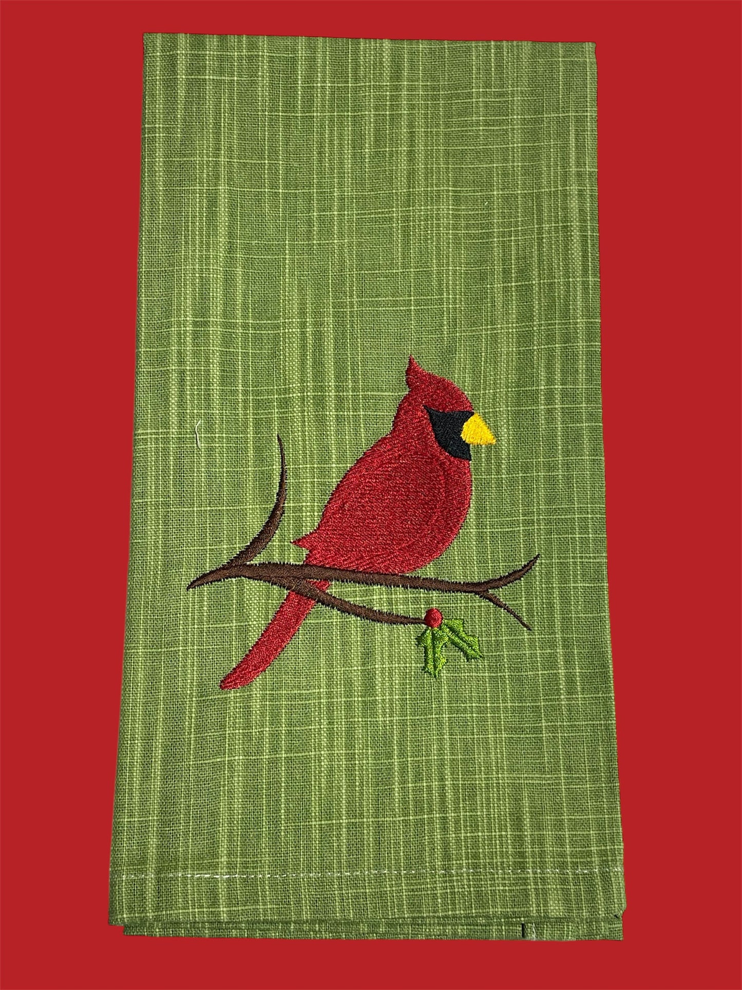 Decorative hand towel featuring embroidered red cardinal sitting on a branch with holly berries, displayed on a green textured background, perfect for Christmas or winter decor.