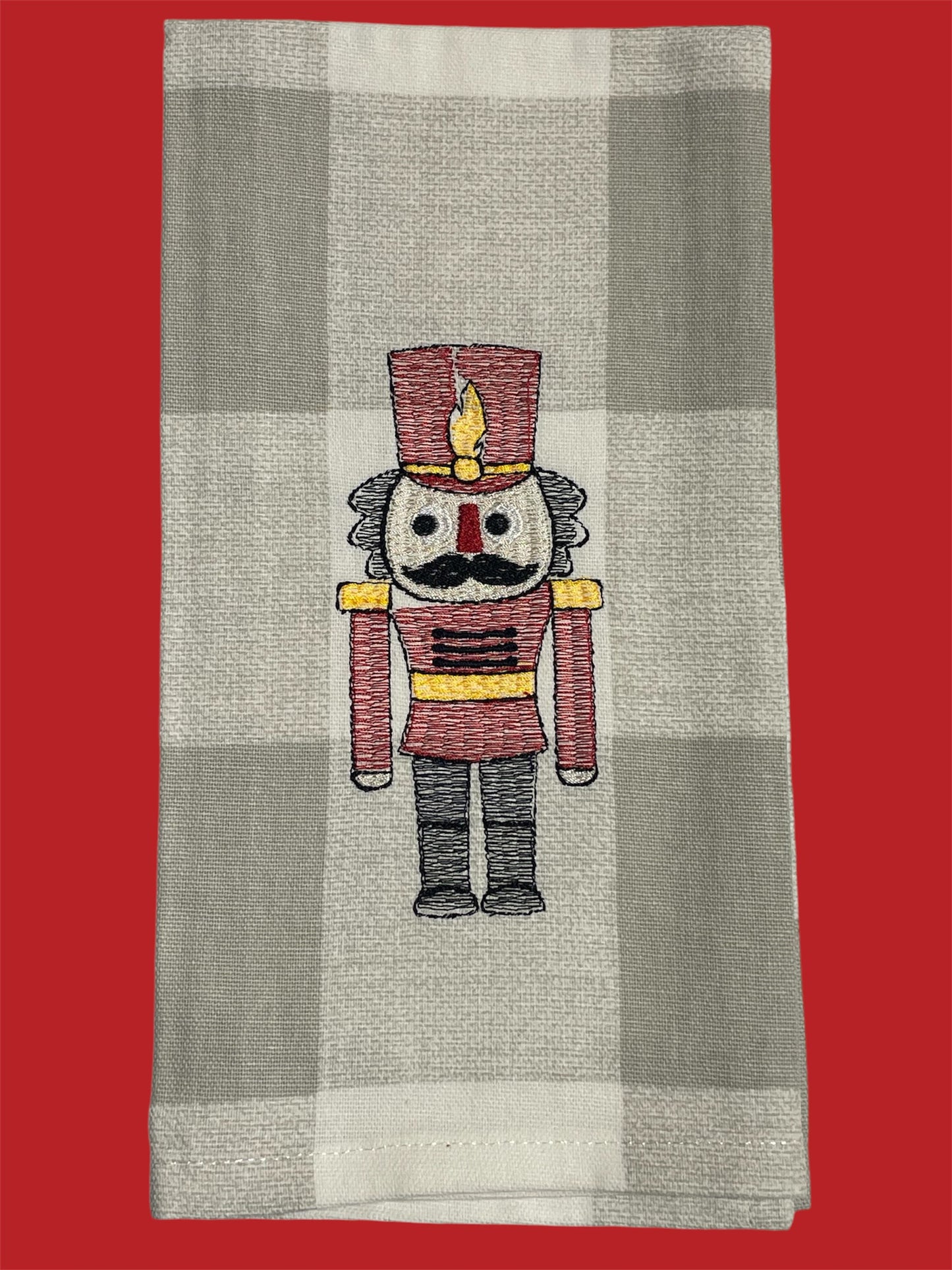 Decorative Christmas hand towel featuring embroidered nutcracker soldier in red uniform with gold and black details on a neutral buffalo check background, perfect for holiday kitchen or guest decor.