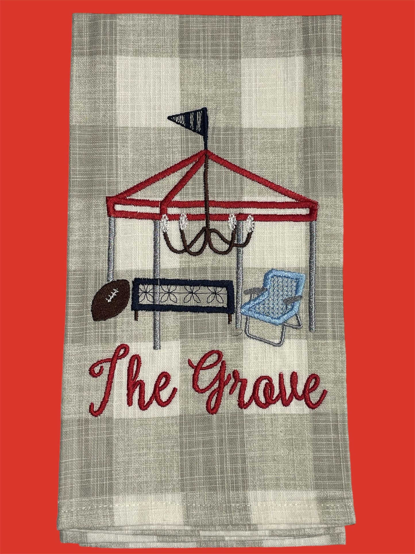 Embroidered hand towel featuring The Grove tailgate scene with tent, chandelier, football, bench, and chair on a light gray plaid background, perfect for Ole Miss fans and Southern tailgating decor.