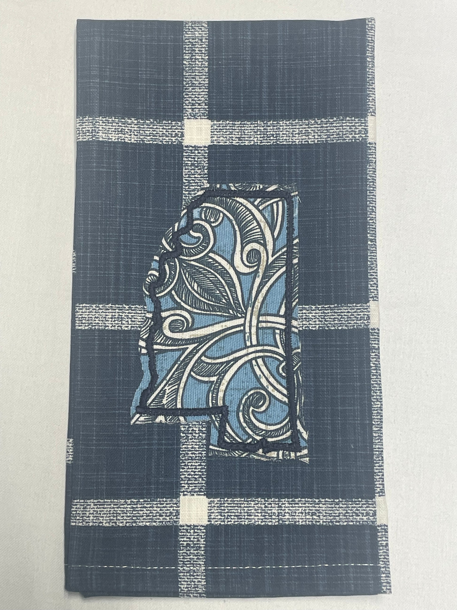 Decorative hand towel featuring embroidered Mississippi state outline with blue and white floral scroll pattern on dark blue plaid background, perfect for southern home decor and state pride gifts.