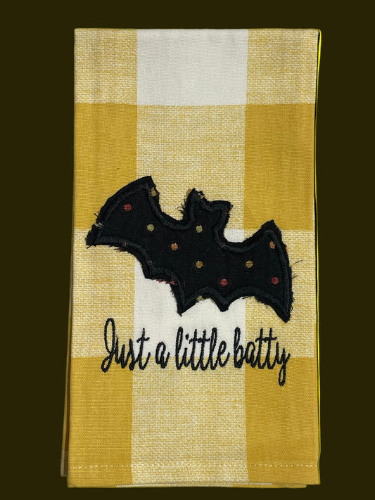 Decorative Halloween hand towel featuring embroidered black bat with polka dots and "Just a little batty" text on yellow gingham background, perfect for seasonal kitchen or party decor.