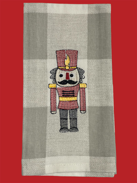 Decorative Christmas hand towel featuring embroidered nutcracker soldier in red uniform with gold and black details on a neutral buffalo check background, perfect for holiday kitchen or guest decor.