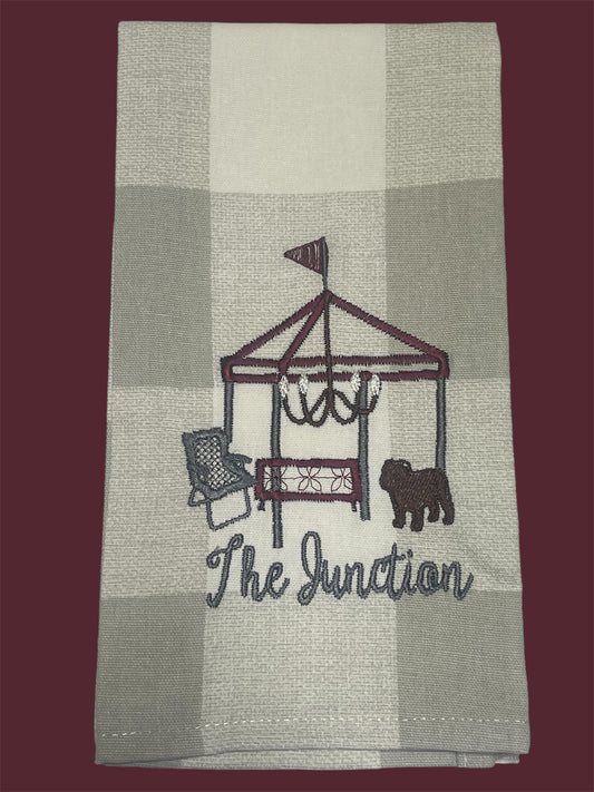Hand towel featuring embroidered tailgate scene with tent, chandelier, chair, bulldog, and "The Junction" text on a neutral buffalo check background, perfect for Mississippi State fans and gameday decor.