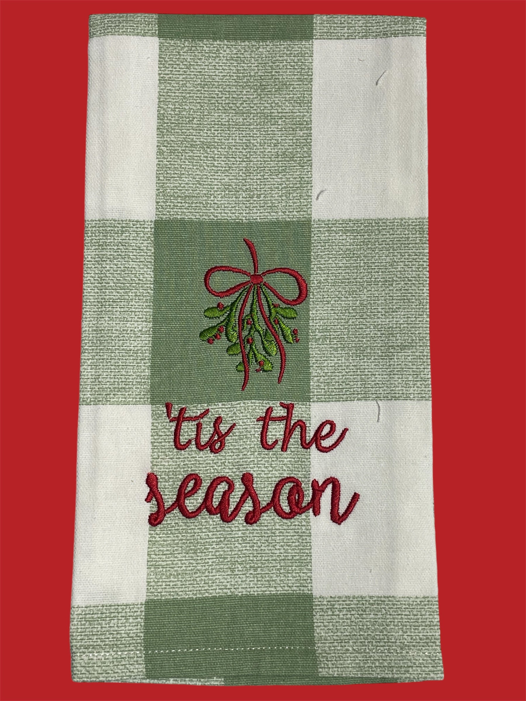 Christmas hand towel with green plaid background featuring embroidered mistletoe with red bow and "’tis the season" text, perfect for holiday decorating or gifting.