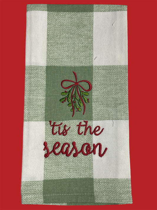 Christmas hand towel with green plaid background featuring embroidered mistletoe with red bow and "’tis the season" text, perfect for holiday decorating or gifting.
